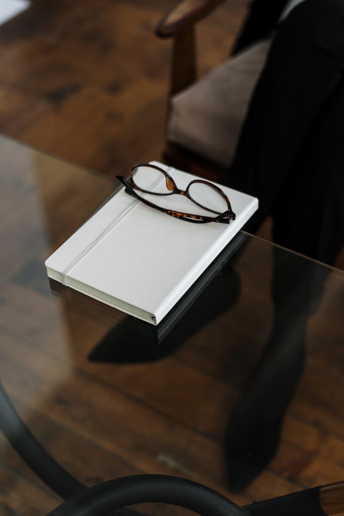 A pair of stylish glasses on a white notebook on a glass table with a wooden floor background.
